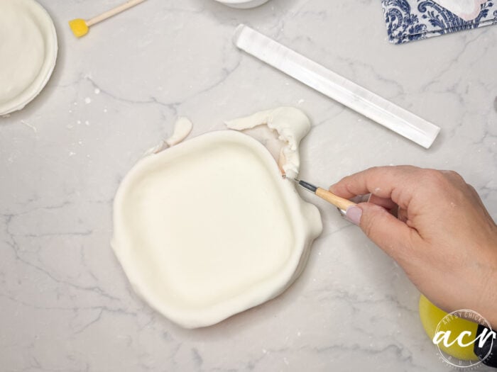 A hand uses a sculpting tool to trim excess diy air dry clay from the edge of a square dish on a marble surface. Tools and materials, including a rolling pin, are nearby.