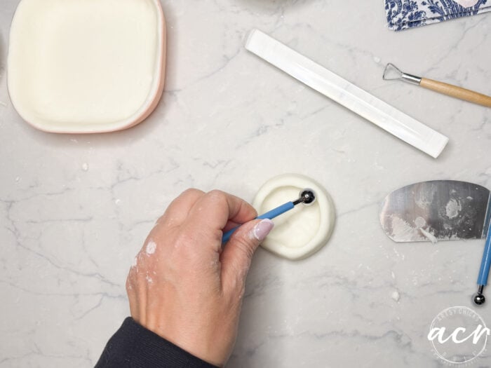 A hand uses a blue sculpting tool to shape DIY air dry clay on a marble surface. Nearby are a soap dish with clay, a white strip, a metal scraper, and another sculpting tool.