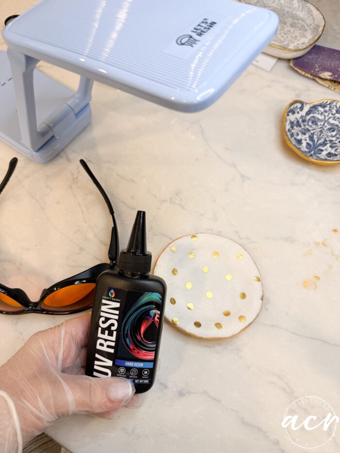 A gloved hand holds a bottle of UV resin above a white circular object with gold dots on a marble surface. Nearby are safety glasses, a UV lamp, and decorative items for making unique decoupage earrings or learning how to use resin safely.