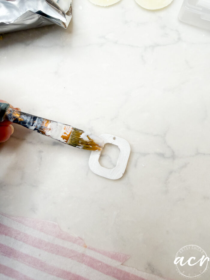 A hand uses a paintbrush to apply white paint to a square-shaped object with rounded edges and two holes, ideal for making decoupage earrings, placed on a light marble surface.