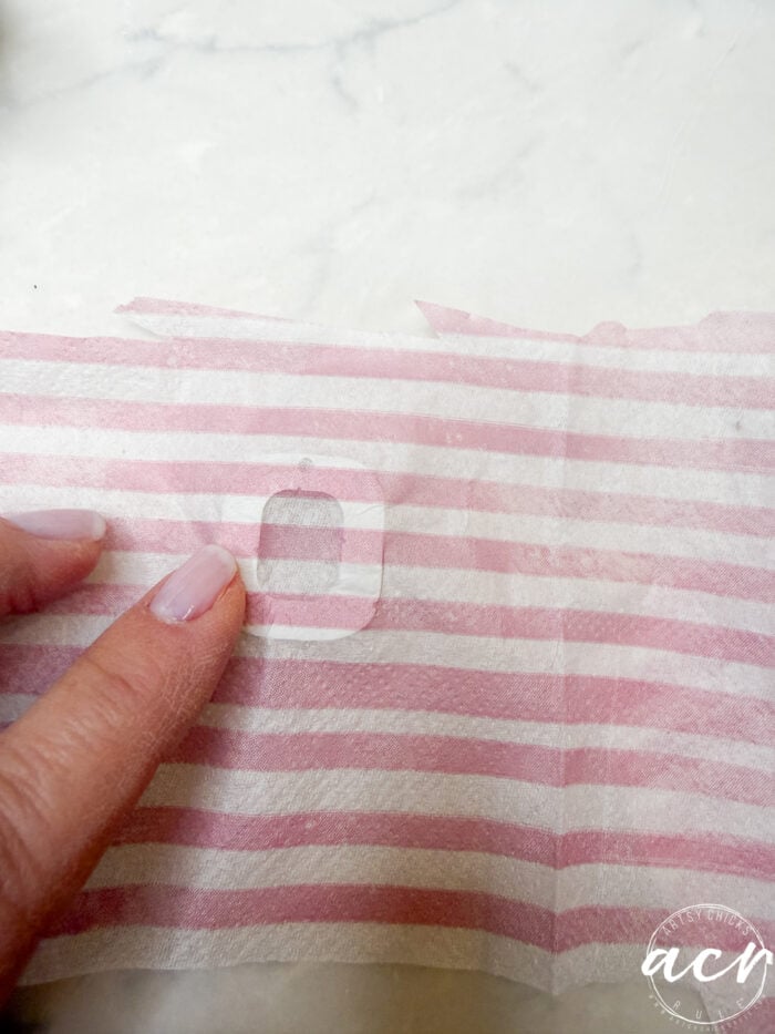 A hand with light pink nails points to a small, clear rectangular sticker on pink and white striped fabric, placed on a light-colored surface. The bottom right corner has a white acy logo—perfect for UV Resin or decoupage earrings projects.
