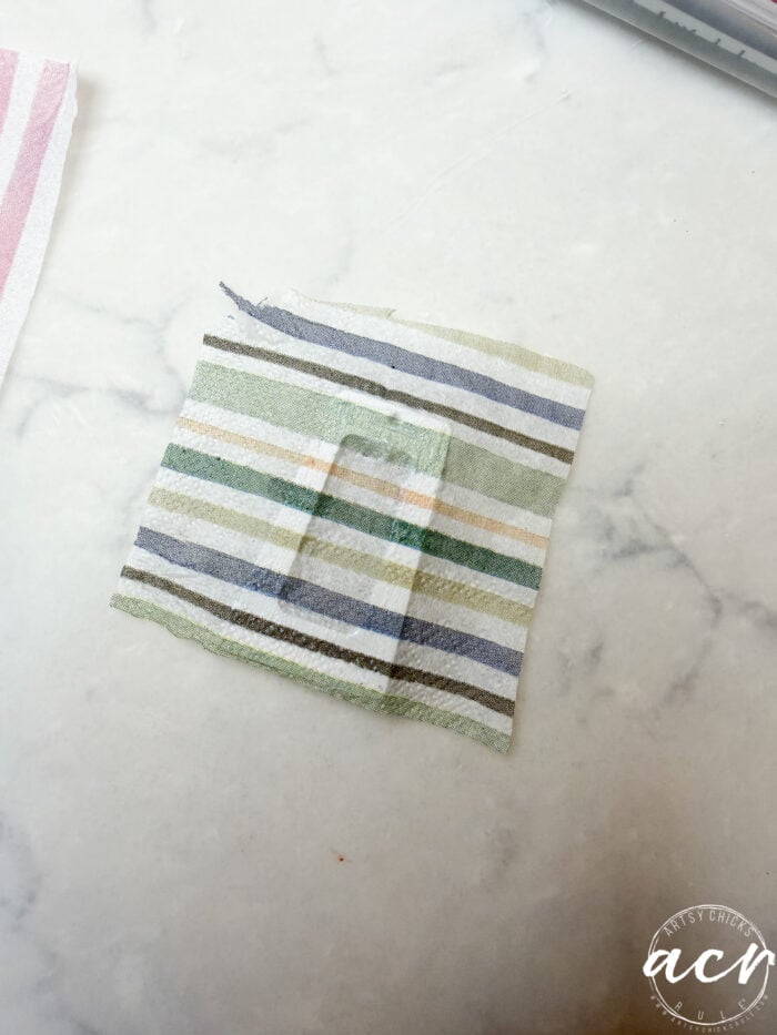 A small square of striped fabric with a rectangular cutout in the center, perfect for decoupage earrings or learning how to use UV resin, is placed on a white marble surface. A logo with the initials ACV appears in the bottom right corner.