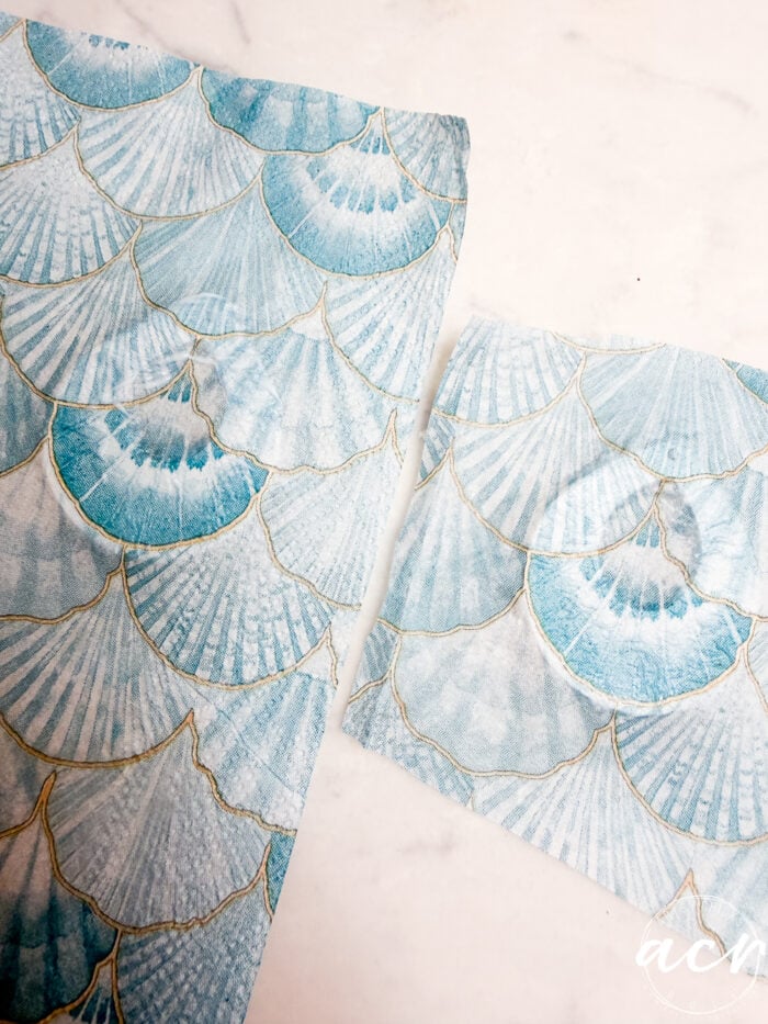 A sheet and a square piece of blue and white paper with a scallop shell pattern outlined in gold, perfect for decoupage earrings or learning how to use UV resin, placed on a white surface.
