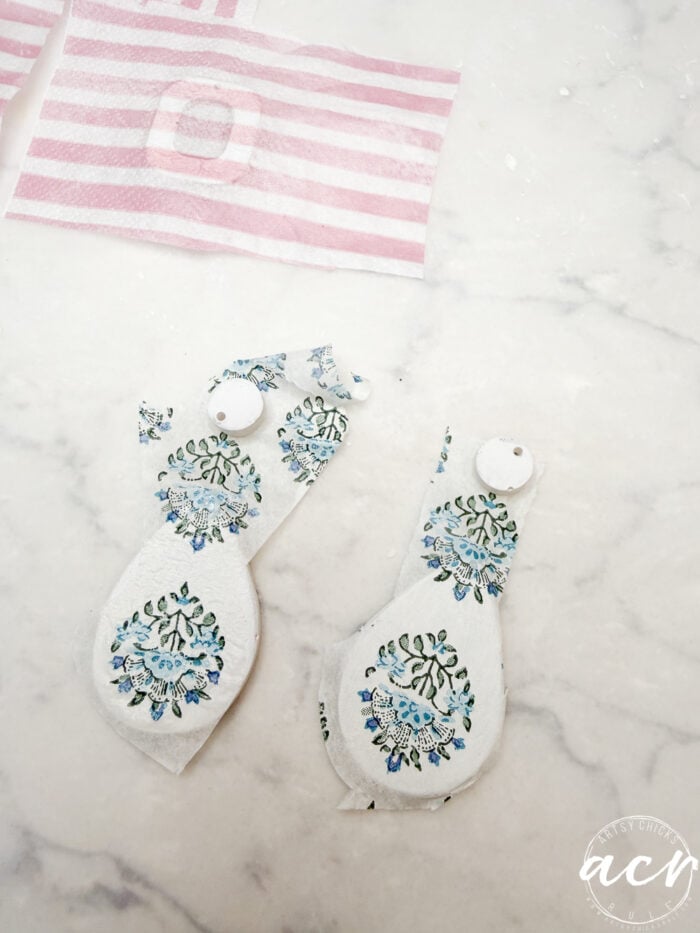 Two pieces of white fabric with blue and green floral patterns cut into ornament shapes lie on a marble surface, perfect for creating decoupage earrings. In the background, pink and white striped fabric pieces await their own UV resin finish.