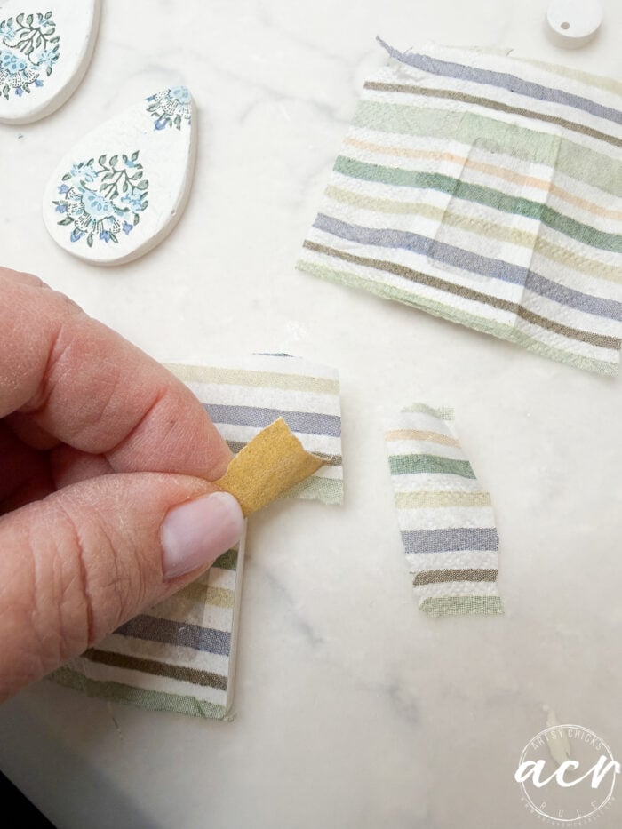 A hand peels striped fabric from a flat surface, with more fabric pieces and decorated white teardrop-shaped decoupage earrings on a white background, illustrating how to use UV resin for crafting.