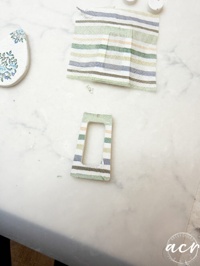 A rectangular piece of striped fabric and a matching, cut-out rectangular earring base—ideal for decoupage earrings or exploring how to use UV resin—rest on a white marble surface. Part of another patterned piece is visible in the corner.