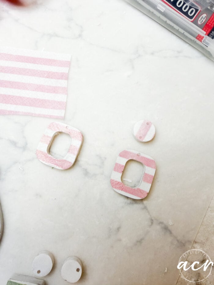 A pair of striped, rectangular and round clay earring pieces in pink and white rest on a marble surface, ready for a coat of UV resin, shown alongside a striped clay sheet and crafting tools.