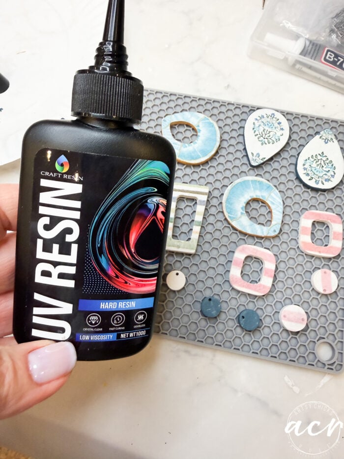 A hand holds a bottle of Craft Resin UV resin above a gray mat filled with colorful decoupage earrings, resin molds, and jewelry pieces. An adhesive tube, additional molds, and finished creations appear in the background.