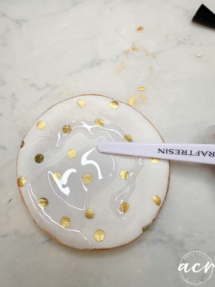 A round white coaster with gold polka dots is being coated with UV resin using a craft stick on a white marble surface, showing how to use resin for a glossy finish.