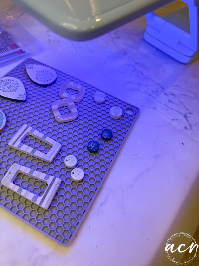 Earrings and earring components, including decoupage earrings, are arranged on a gray silicone mat under a blue crafting lamp, with a marble surface beneath. Some pieces are white, while others feature dark blue with white speckles and hints of UV resin.