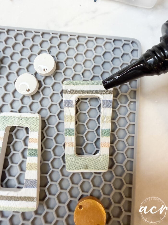 A hand applies glue to a rectangular, striped fabric pendant on a gray hexagonal mat, with circular and round components nearby—perfect for crafting decoupage earrings or learning how to use UV resin.