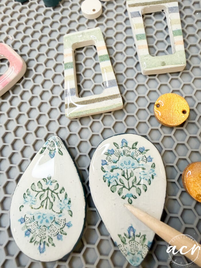 Handmade ceramic jewelry pieces on a hexagonal mat, featuring decoupage earrings in teardrop shapes with blue floral designs, striped rectangular frames, and round orange pendants; a wooden stick points at a teardrop piece.