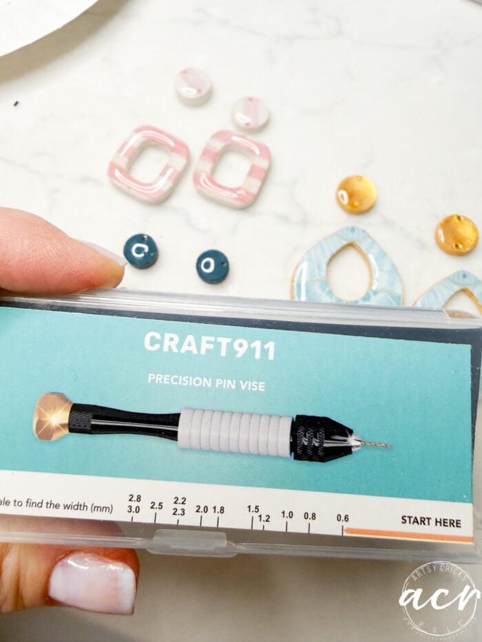 A hand holds a CRAFT911 Precision Pin Vise tool in its packaging, with colorful decoupage earrings, UV resin supplies, and jewelry-making beads artfully arranged on a white surface in the background.