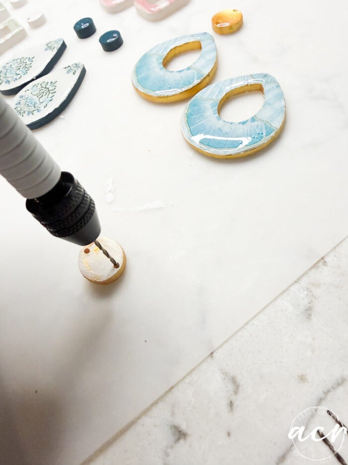 A person uses a power drill to make a hole in a round piece of blue and tan polymer clay, with various UV resin-coated clay earring pieces on a white surface nearby.