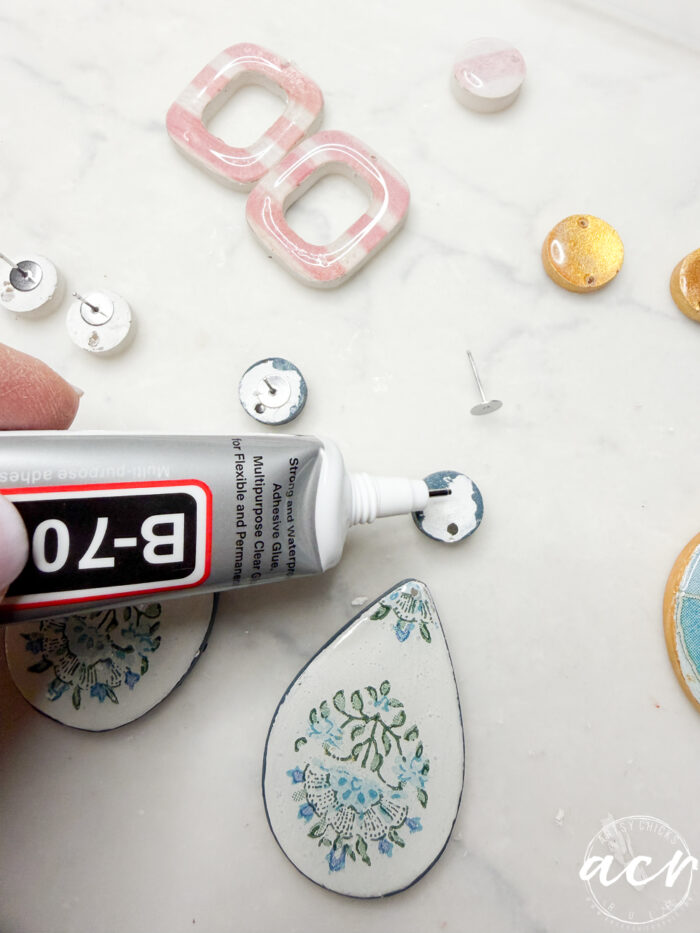 A hand applies B-7000 adhesive to the back of a round earring post; various colorful decoupage earrings and posts are arranged on a white surface, illustrating how to use different materials in jewelry making.