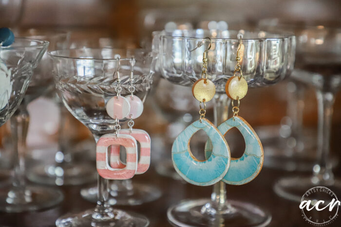 Two pairs of colorful decoupage earrings hang from the rims of glass goblets; one pair is pink and white with geometric shapes, the other blue and gold with a teardrop design. The background features more glassware.