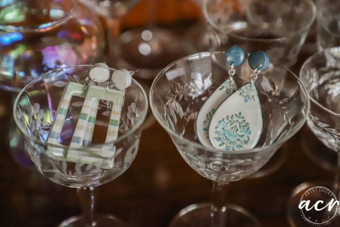 Two pairs of decoupage earrings displayed inside vintage etched glass goblets: one pair with green and white stripes, the other with blue and white teardrop shapes. The background features more glassware and soft reflections.