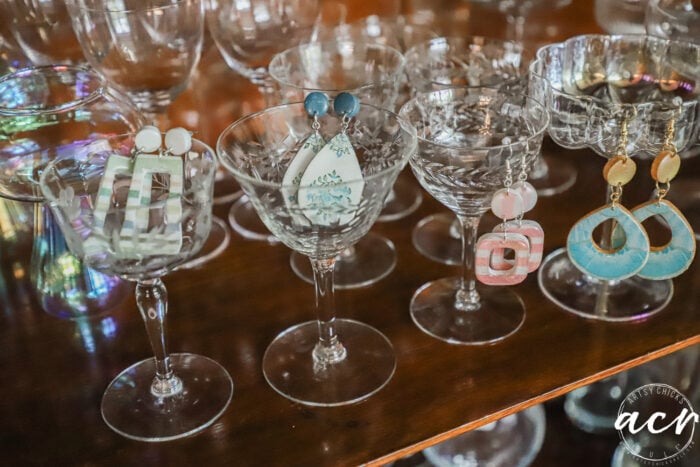 Several pairs of colorful, geometric decoupage earrings are displayed hanging on the rims of clear cocktail glasses, which are arranged on a wooden surface. The background shows more glassware, and the image is signed acv.