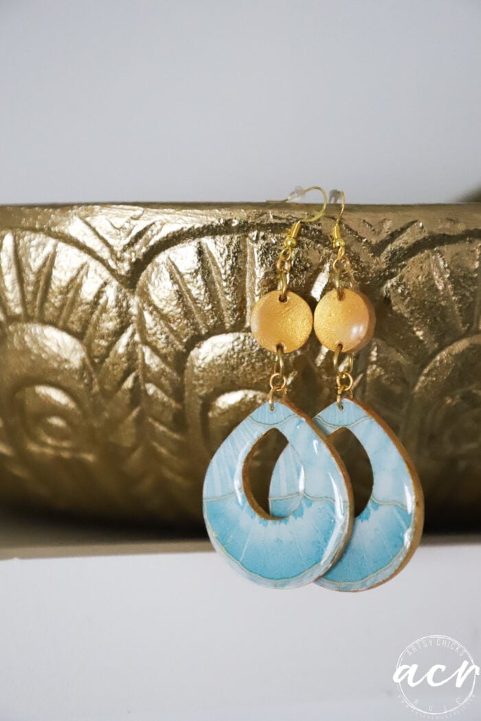 A pair of handmade decoupage earrings with gold round beads and large, oval-shaped blue UV resin drops hang in front of a textured gold decorative surface.