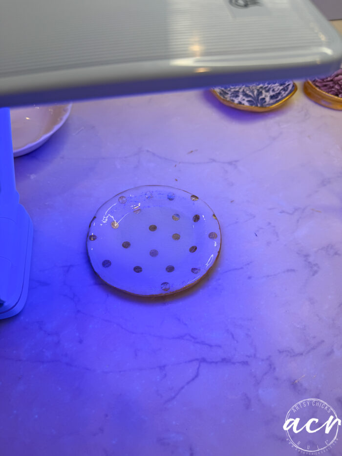 A small round dish with gold polka dots is placed under a UV lamp on a white marble surface, likely curing UV resin. Other decorative dishes, possibly for decoupage earrings, are visible in the background, suggesting a creative crafting process.