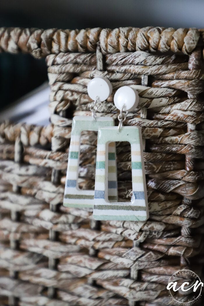 A pair of rectangular, striped decoupage earrings with white circular studs are hanging on the woven surface of a rustic basket.