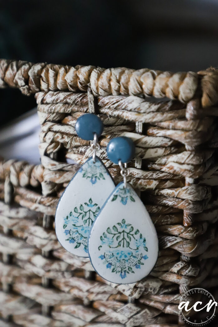 A pair of decoupage earrings featuring teardrop shapes with blue and green floral designs and blue round tops, hanging on a woven basket. Perfect for showcasing unique style or learning how to use UV resin in jewelry making.