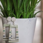 A pair of geometric, striped decoupage earrings hang from the rim of a white flower pot with green, spiky leaves. The pot has a faint blue smudge and a white logo in the bottom right corner, showcasing how to use creative techniques.