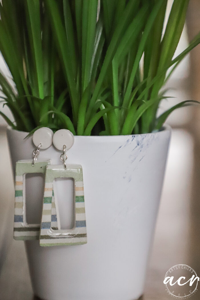 A pair of geometric, striped decoupage earrings hang from the rim of a white flower pot with green, spiky leaves. The pot has a faint blue smudge and a white logo in the bottom right corner, showcasing how to use creative techniques.
