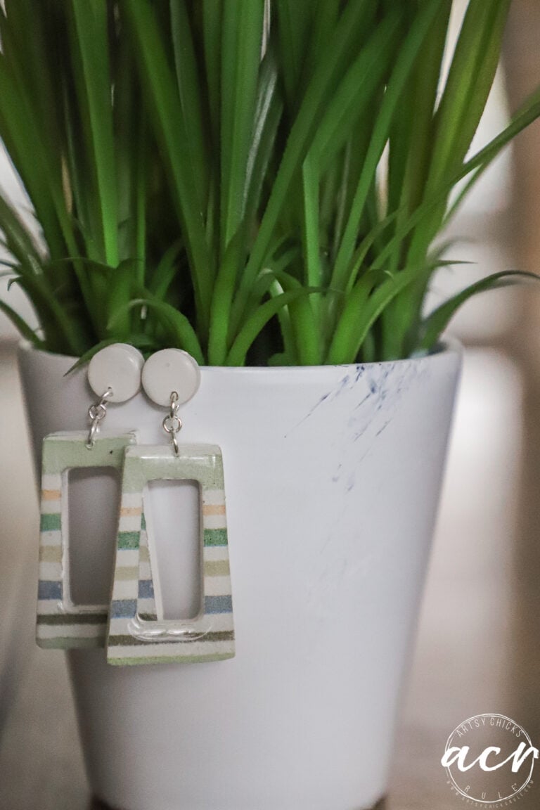 A pair of geometric, striped decoupage earrings hang from the rim of a white flower pot with green, spiky leaves. The pot has a faint blue smudge and a white logo in the bottom right corner, showcasing how to use creative techniques.