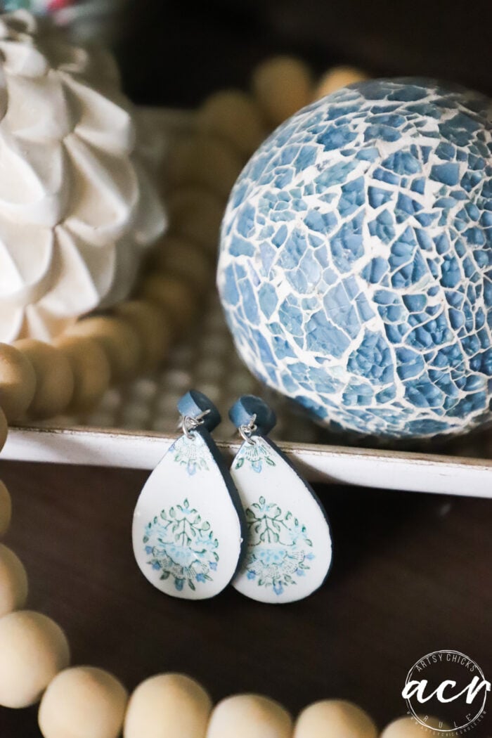 A pair of teardrop-shaped decoupage earrings with blue and green floral designs are displayed in front of a blue crackle-patterned decorative sphere and a white textured object, with wooden beads nearby. Perfect for those learning how to use UV resin.