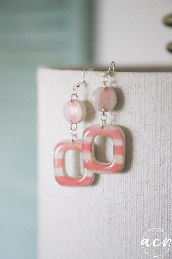 A pair of handmade decoupage earrings with pink round beads and square, pink-striped frames hang from a textured beige fabric background. The soft lighting enhances their artistic style, perfect for learning how to use UV resin in your own creations.