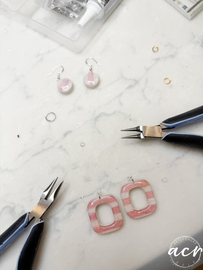 A marble surface with jewelry-making tools, decoupage earrings in pink and white, jump rings, and a small open organizer with supplies. The scene suggests a DIY jewelry crafting project, perfect for exploring how to use UV resin.