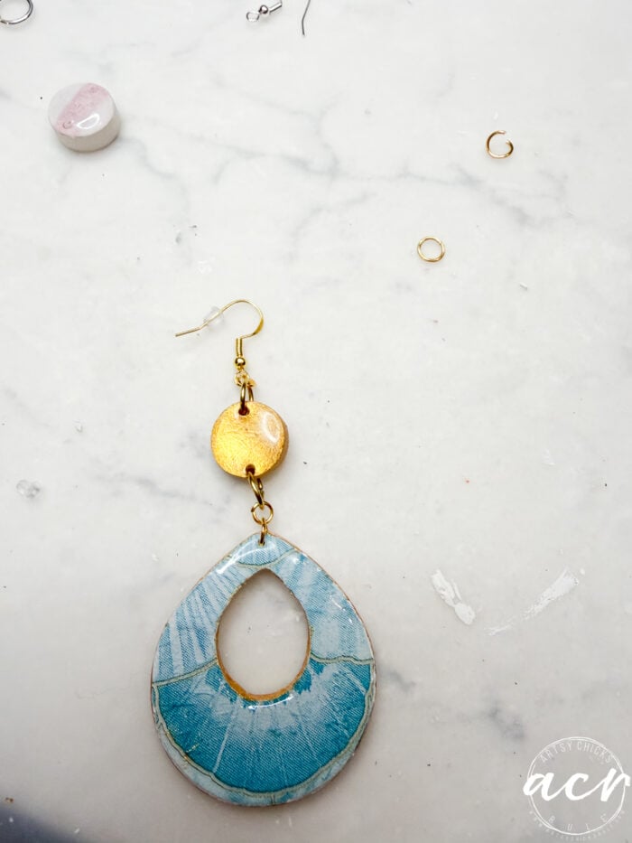 A handmade Decoupage Earring with a blue teardrop-shaped bottom and a small round gold piece above it rests on a white marble surface, surrounded by a few small jewelry-making components, perfect for learning How To Use UV Resin.