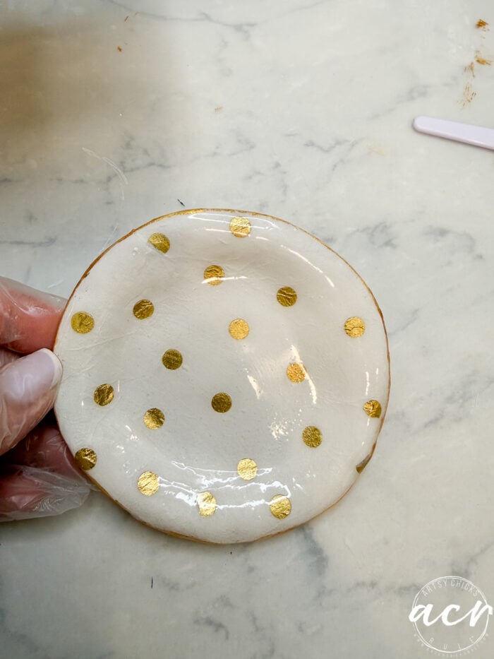 A hand wearing a glove holds a small, round white dish with shiny gold polka dots—perfect for displaying decoupage earrings or curing creations in UV resin—set against a white marble surface.