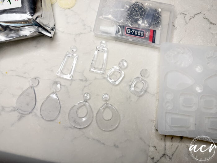 Clear resin earring pieces in various shapes are laid out on a marble surface. Nearby are a silicone mold, UV Resin, B-7000 glue, earring hooks, and a partially visible metallic pouch—perfect for learning how to use decoupage earrings.