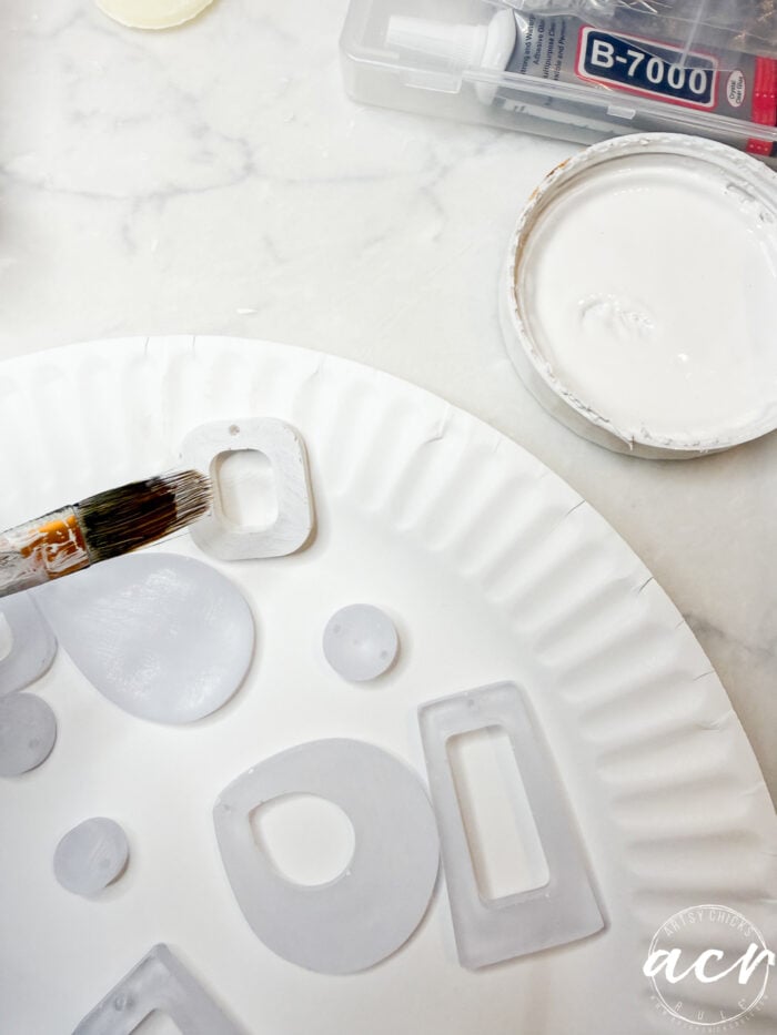 A paper plate holds translucent decoupage earrings being coated with UV resin by a paintbrush. Nearby are an open container of white resin and a tube of B-7000 adhesive on a marble surface, showing how to use these materials for crafting.
