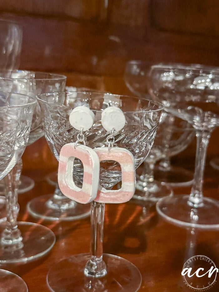 A pair of pink and white square-shaped decoupage earrings with round tops are displayed on the rim of a vintage cut-glass goblet, surrounded by other similar glassware on a wooden surface.
