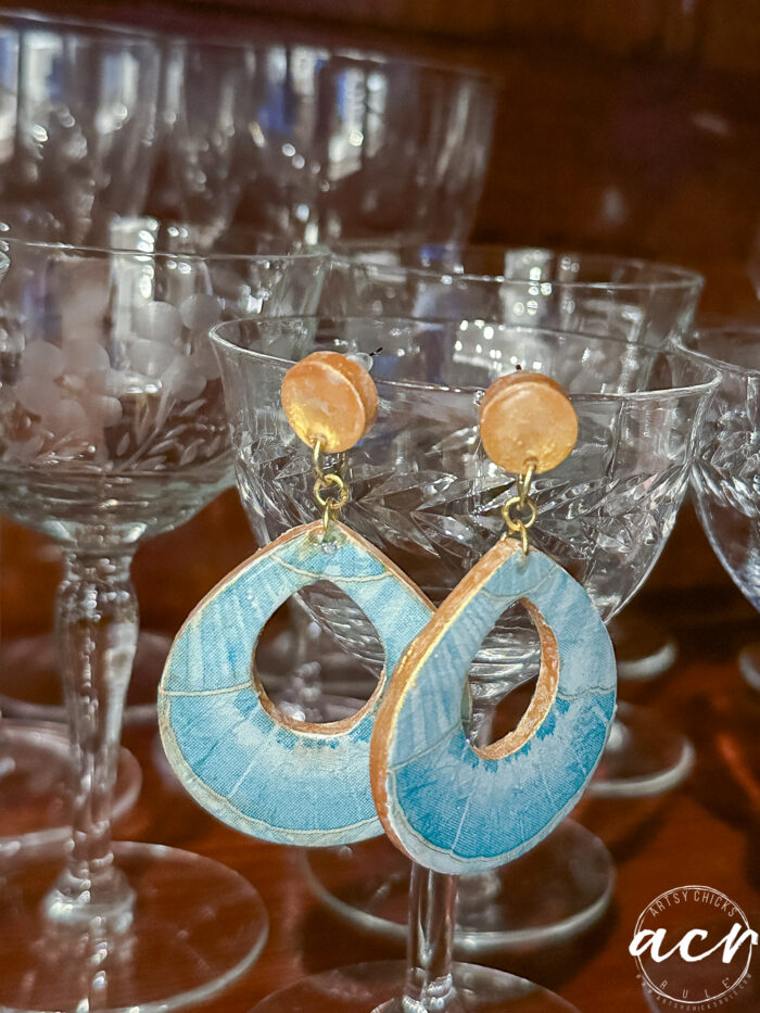 A pair of blue and gold decoupage earrings with round tops and large teardrop-shaped bottoms, displayed on the rim of clear glass goblets. The background features more stacked glassware—showcasing how to use UV resin for a glossy finish.