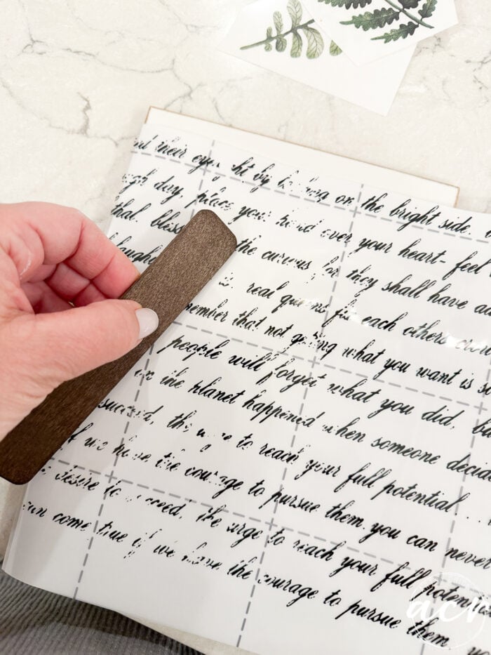 A hand uses a brown tool to rub black script lettering onto a white sheet, with Easter Edition plant stickers nearby on a marble surface.