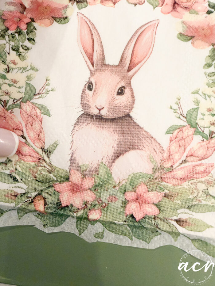 Illustration of a brown rabbit with pink ears sitting among pink flowers and green leaves, framed in a floral wreath on a green background. A hand with light-colored nail polish is visible on the left side&mdash;perfect for an Easter Edition of Flipping Smalls.