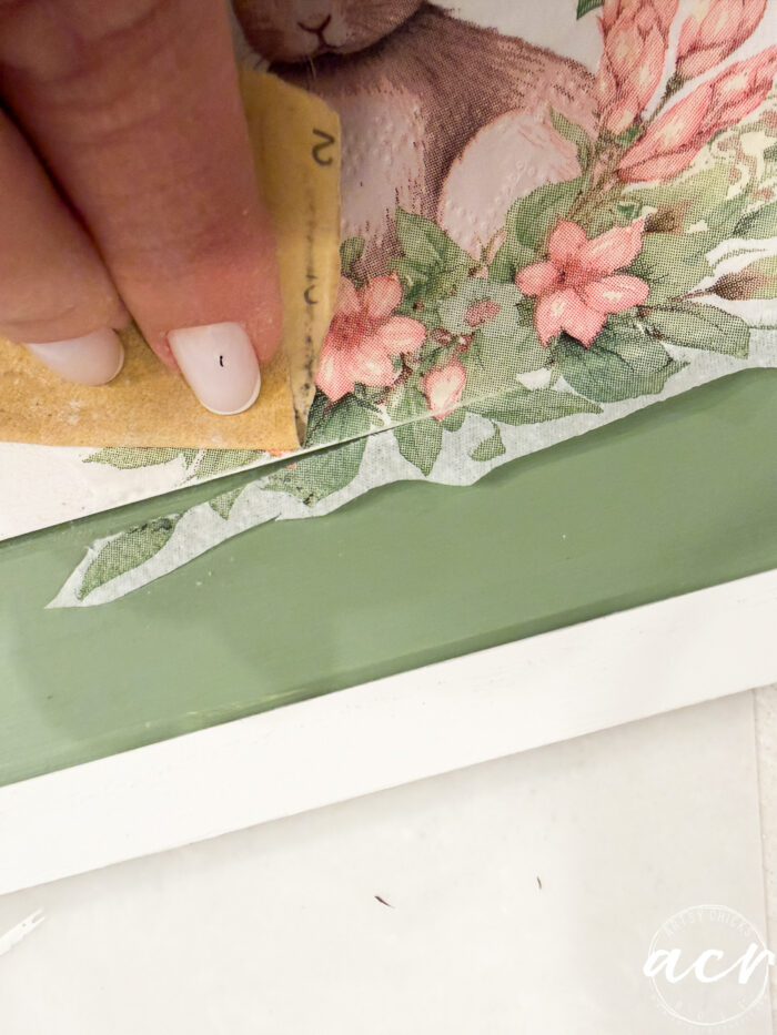 A hand with light nail polish uses sandpaper to smooth the edge of a floral and bunny illustration on a green and white surface, creating an adorable Flipping Smalls piece for the Easter Edition.
