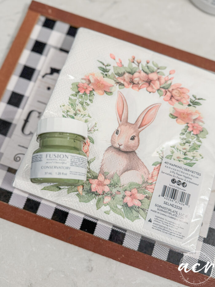 A small jar of green Fusion Mineral Paint, part of the Easter Edition Flipping Smalls, sits on a tray next to napkins featuring a rabbit in a floral wreath. The background includes a black and white checkered pattern.