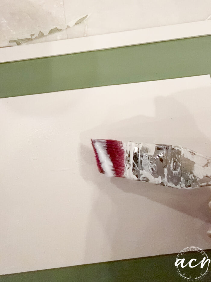 A paintbrush with white paint is being applied to a flat white surface in this Easter Edition image. The background shows a green and white area, and a watermark with the text &ldquo;acv&rdquo; appears in the bottom right corner.