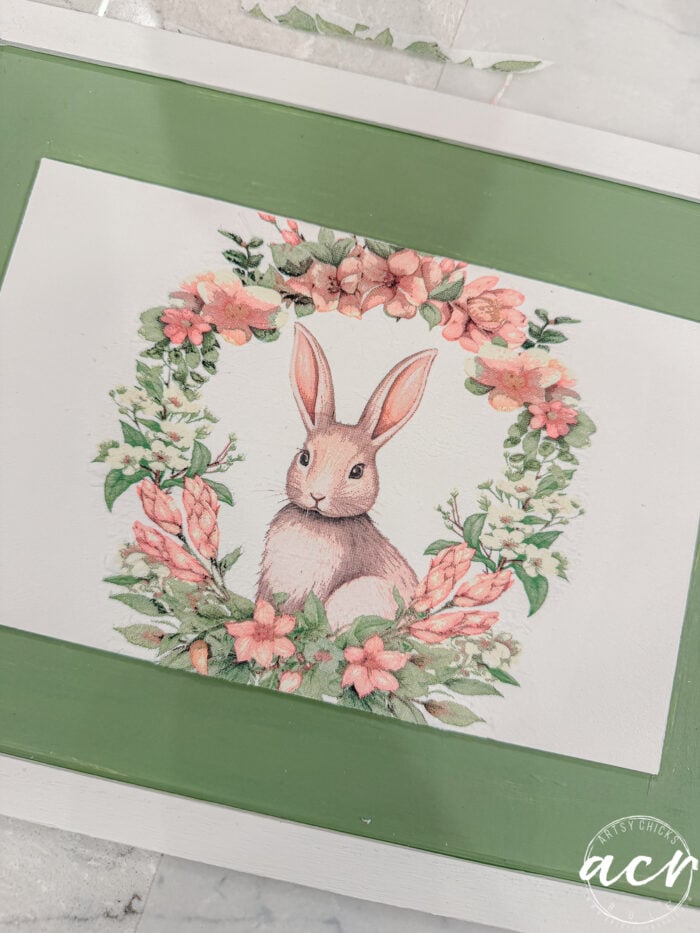 A painted illustration of a brown bunny sitting inside a wreath of pink and green flowers, bordered by a green rectangular frame. This Easter Edition image has a soft, pastel aesthetic perfect for Flipping Smalls collections.