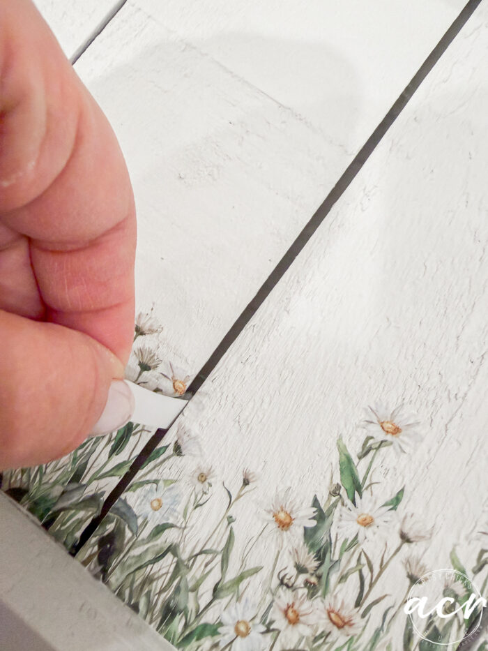 A hand peels up a strip of decorative floral transfer from a white wooden surface, revealing a painted design of green leaves and small white flowers&mdash;perfect for your Flipping Smalls Easter Edition craft projects.