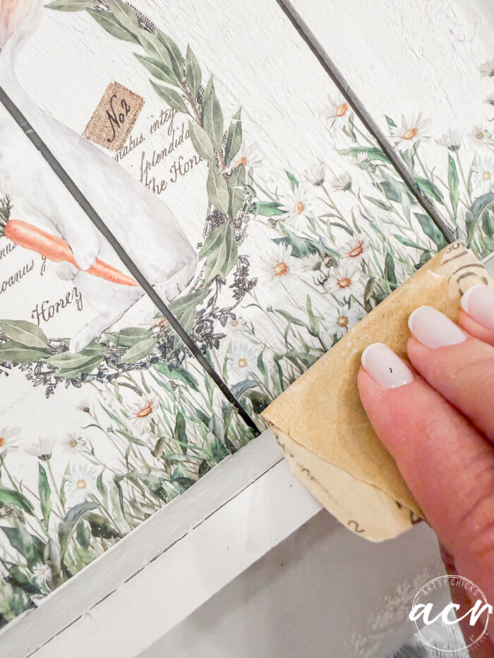 A hand with neatly manicured nails uses sandpaper to distress the edge of a painted wooden surface decorated with floral and greenery designs&mdash;perfect for your Flipping Smalls Easter Edition projects.