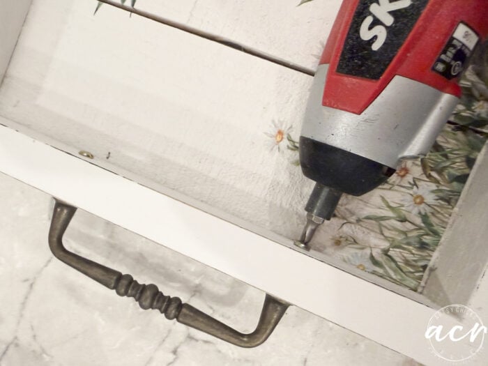 A close-up of a red power drill driving a screw into a white wooden tray with a floral design and metal handle, perfect for your Flipping Smalls Easter Edition DIY project. The tray rests on a light-colored marble surface.