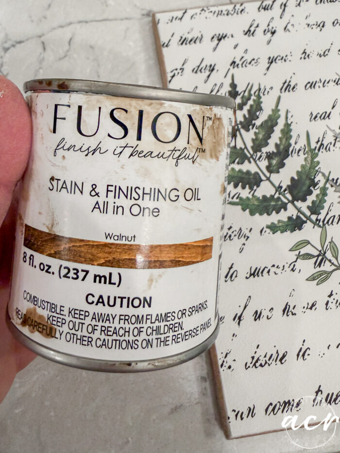 A hand holds a can of Fusion Stain & Finishing Oil (Walnut, 8 fl. oz.) in front of a decorative art piece with green botanical illustrations and script text&mdash;perfect for Flipping Smalls or creating an Easter Edition masterpiece. The can displays cautionary information.
