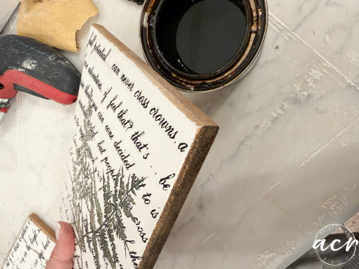 A hand holds a wooden sign with cursive writing and a leaf illustration near a cup of dark stain, a rag, and a stapler on a light work surface. The freshly stained edge adds charm to this Flipping Smalls Easter Edition piece.