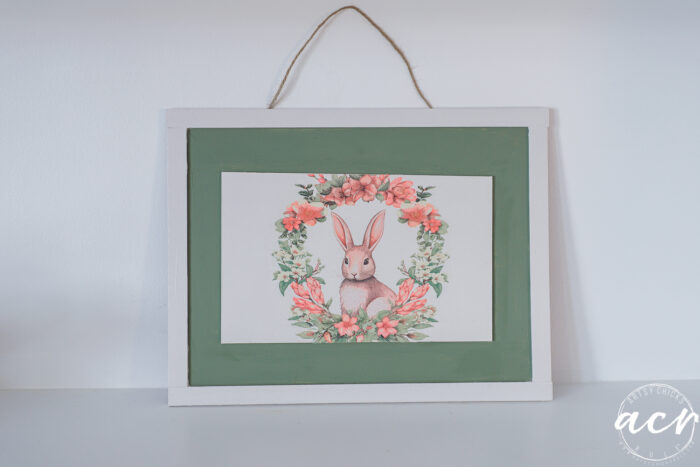 A framed picture of a brown rabbit surrounded by pink flowers and green leaves, hanging on a wall with a twine string. The frame is white with a green inner border, perfect for the Flipping Smalls Easter Edition collection.